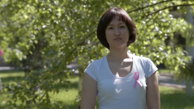 Young brunnete woman in white t-shirt wearing pink ribbon, trees background. Breast cancer awareness