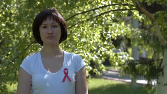 Young woman in white t-shirt wearing red ribbon, green nature background. AIDS and HIV awareness, heart disease, lymphoma, tuberculosis, substance-abuse, vasculitis