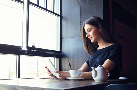 Young attractive girl sitting alone near big window in coffee shop during free time and working on tablet computer. Happy female having rest in cafe. Lifestyle, coffee