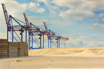 Fototapeta na wymiar tall cranes streydery, cranes and concrete loads in the desert next to the sandy terrain. cargo operations. Sky with krasivmyi clouds in the background 