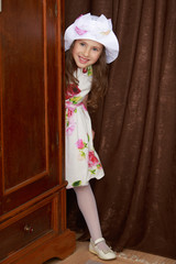 Cheerful little girl in a smart white dress with the roses peeping from behind the old Cabinet