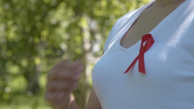 Close-up shot of woman pinning red ribbon on white t-shirt, green nature background. AIDS and HIV awareness, heart disease, lymphoma, tuberculosis, substance-abuse, vasculitis