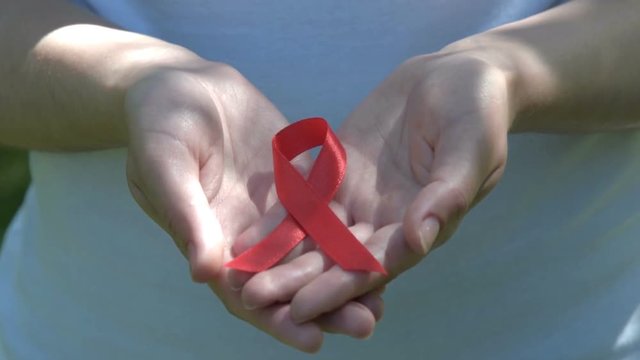 Close-up shot of woman hands holding red ribbon in sunlight. AIDS and HIV awareness, heart disease, lymphoma, tuberculosis, substance-abuse, vasculitis
