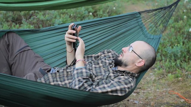 A man in a hammock touch phone. Man in the woods with a beard and glasses