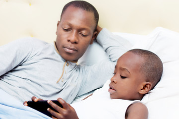 Fototapeta premium Dad looks at his child using mobile phone in bed