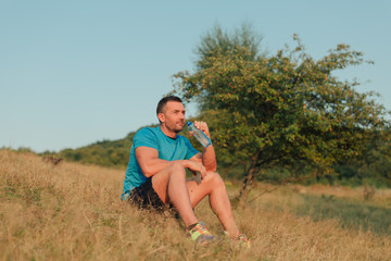 Naklejka na ściany i meble Athletic man in his 30s resting and drinking water from a bottle while sitting down outdoor