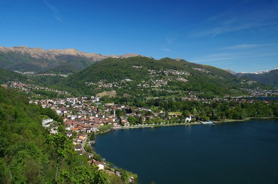 Swiss city of Caslano at the west side of the Lago di Lugano. Bare and snow covered mountains in the background. Densely forested hills spotted with houses steeply descend to the lake shore.