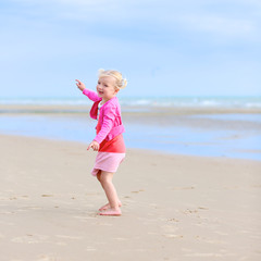 Cute active child wearing pink dress playing and dancing on wide sandy beach. Happy little girl enjoying summer holidays on a sunny day. Family with young kids on vacation at the North Sea coast.