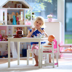 Cute preschooler girl having birthday tea party with her doll. Little child plays in sunny room at home or kindergarten. Kids using toy dishes and cakes.