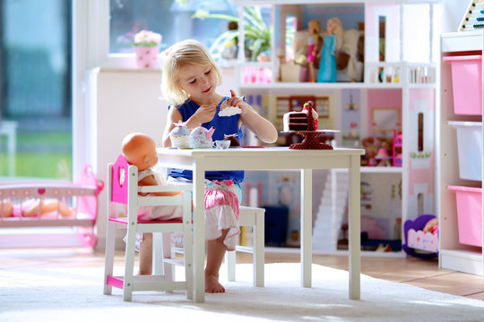 Cute Preschooler Girl Having Birthday Tea Party With Her Doll. Little Child Plays In Sunny Room At Home Or Kindergarten. Kids Using Toy Dishes And Cakes.