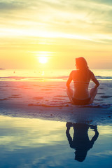 Young woman sitting on the beach during sunset, silhouette reflection in water.