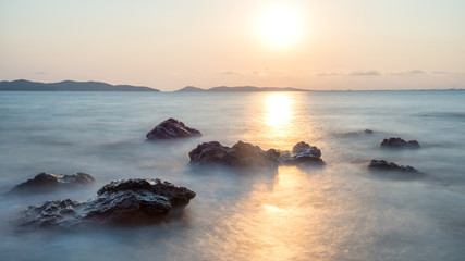 Fototapeta na wymiar Romantic atmosphere in peaceful sunset at sea. Big boulders sticking out from smooth wavy sea.