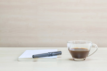 Closeup black pen on white book with black coffee in transparent cup of coffee on blurred wooden desk and wall textured background in the meeting room under window