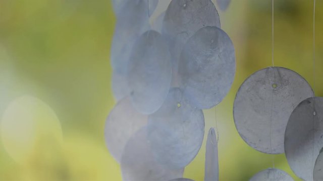 Macro closeup of moving capiz shell wind chimes against bokeh sunlight garden background, handheld, with light leaks.
