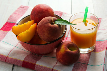 delicious and juicy peaches with green leaves and chopped peach pieces in a bowl on white wooden background near the glass of peach juice on a napkin