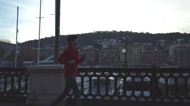 Female jogging in town. Young runner training on the street along road. Urban view with buildings on the background. Athlete runs in red sports sweatshirt with long- sleeved grey tights and sneakers