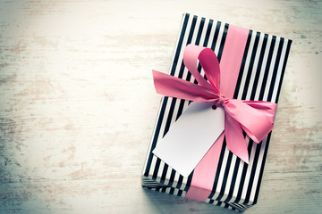 Gift box wrapped in black and white striped paper with pink ribbon on a white wood old background. Empty note tied over. Vintage style.