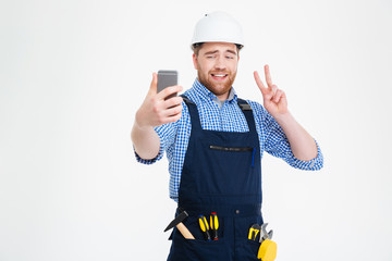 Smiling builder showing peace sign and making selfie with smartphone