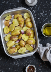 Raw potatoes with olive oil and spices in the baking dish. The preparation of baked potatoes. On a dark stone background.