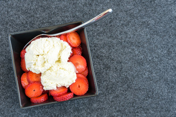 Sliced strawberries and vanilla ice cream with a spoon in a black square bowl on a black background 

