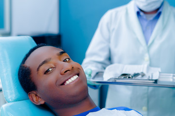 Young hispanic male lying in dental chair looking to camera smiling, dentist coat visible background