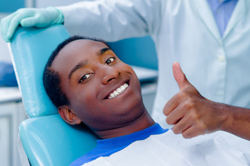 Young hispanic male lying in dental chair looking to camera smiling giving thumb up, dentist coat visible background