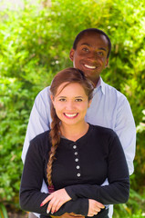 Beautiful young interracial couple in garden environment, embracing and smiling happily to camera