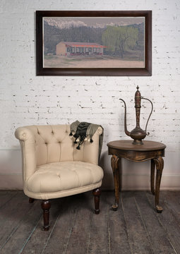 Vintage still life of  old beige armchair,  brass teapot, framed painting and small antique table on dark brown wooden floor and white bricks wall in Studio