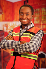 Young carpenter worker wearing square pattern flanel shirt and red safety vest, posing with arms...