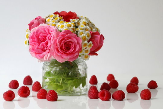 Summer cheerful bouquet of roses and daisies flowers and raspberries. Floral still life with bouquet of pink roses and white mini chrysanthemums in a vase and ripe raspberries. Flower home decoration.