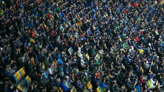 Football supporters wearing warm clothes, sitting on tribunes and watching match