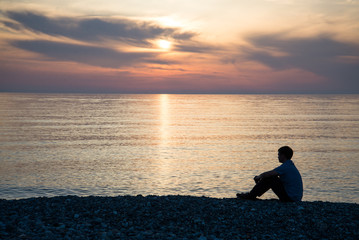Naklejka na ściany i meble Man silhouette sitting on beach with sea and sunset background