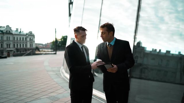 Two young europian businessmen have a deal and communicate. First with tablet PC making offer to second. Second with coffee reject his presentation. Glass business centre building at the background