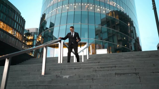 Young successful businessman easy sliding on the outdoor stairs rail with smile and jump at the end. Glossy business centre building at the background. Teal and Orange style. Wide 4k tripod shot.