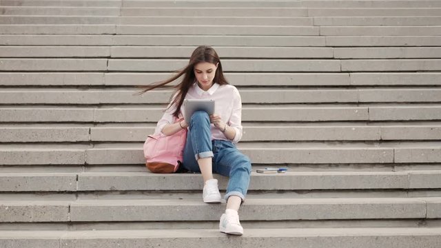 Young beautiful stylish girl sits on steps and holding hands in the tablet. Girl student communicates in social networks with friends.