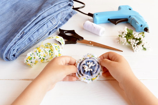 Child holds brooch denim flower in his hands. Creative flower brooch using old jeans. Handmade recycled denim jeans accessory. Kids fabric art. Scissors, glue gun, lace, thread, needle on a wood table