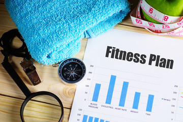 "Fitness Plan" text on paper with blue bar chart on wooden table with compass,magnifying glass, pen, towel, green apple with measurement tape, and whistles - fitness, diet and healthy concept