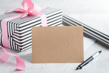 Empty greeting kraft card with pen. Wrapped gift and wrapping paper roll in the background.