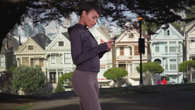 Sporty black woman using smartphone texting app in park. African American millennial girl with cell phone standing in front of Painted Ladies of San Francisco.