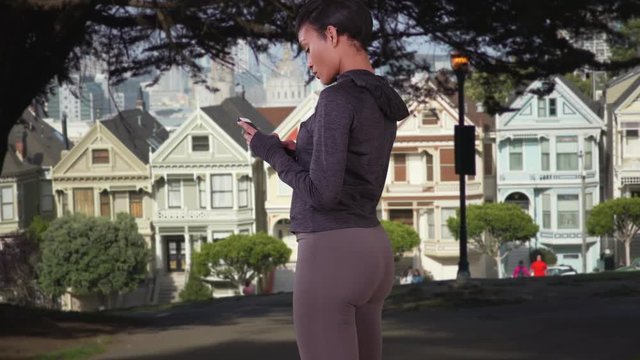 Sporty black woman using smart phone texting app in park. African American millennial girl with cellphone standing in front of Painted Ladies of San Francisco.
