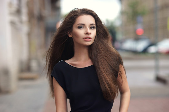 Fashion style portrait of young beautiful elegant woman in black dress walking at city streets on a windy dull day.