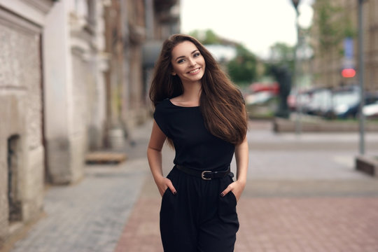 Fashion style portrait of young happy smiling beautiful elegant woman in black dress walking at city streets.
