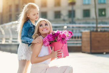 Portrait of young beautiful mother and daughter with box of peonies posing in the city streets. Flower box and gift box. Bouquet of flowers in gift box. Birthday. March 8