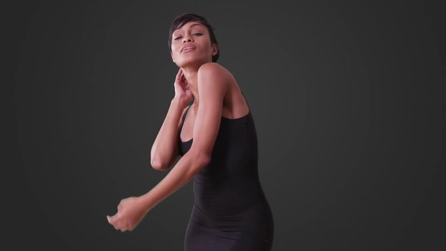 Sexy woman in tight black dress dancing on grey background. African American woman smiles and looks at camera.