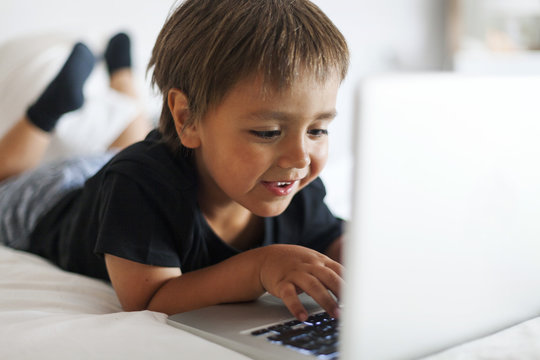 Smiling little boy lying on bed using laptop