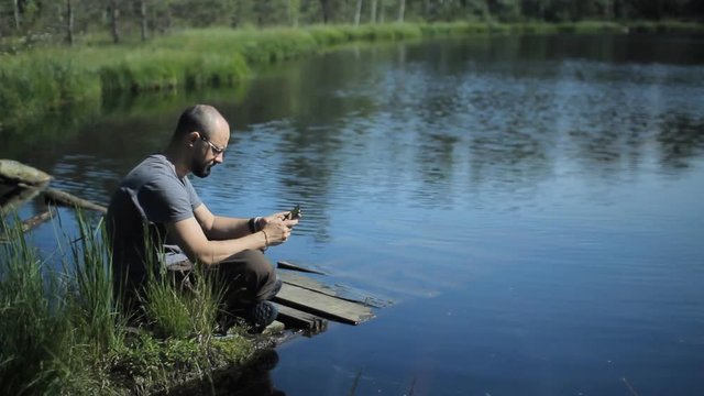 A man sits on a pier of the lake and touch tablet. The beautiful blue lake and forest on background