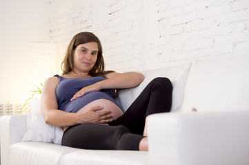 young beautiful happy 8 or 9 months pregnant woman at home living room couch holding big belly