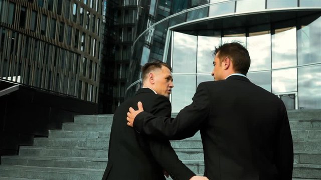 Slow motion. Colleagues and friends. Two cheerful businessmen pat on the shoulder and talking to each other while walking stairs outdoors. Glass Business centre bg.
