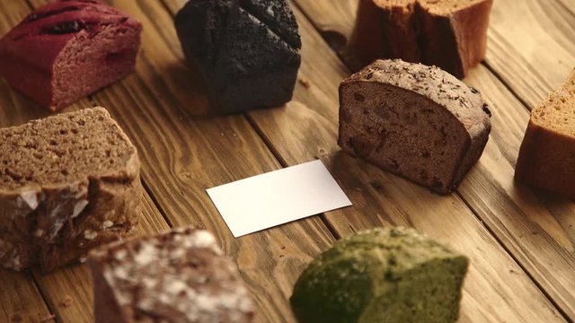 Tattooed hand puts blank business card in center of rustic table with many samples of artisan breads made from beetroot,lavender,potato,spinach,tomatoes,rye and others Professional bakery presentation