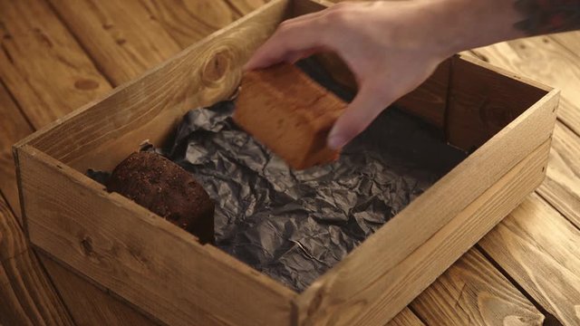 Baker with tattooed hand carefully puts different samples of mixed healthy breads from vegetable and fruits inside rustic wooden box with craft paper on old brushed table for presentation.
